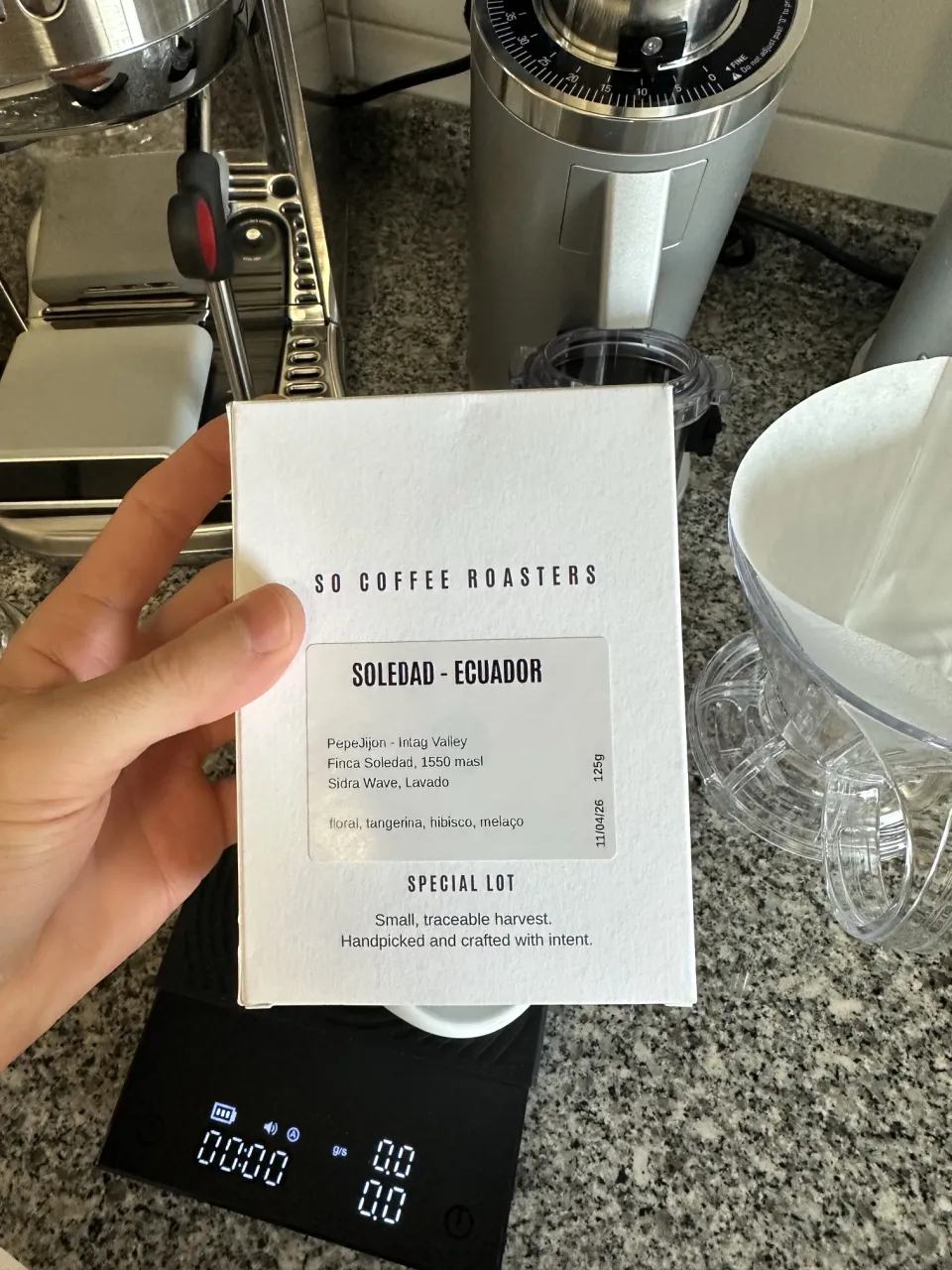 André holding a box of SO Coffee Roasters Soledad, Ecuador, in front of his espresso machine and grinder